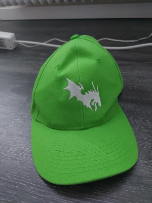 Grön keps med drakmotiv - Snygg grön keps med böjd skärm och justerbar rem bak. På framsidan finns ett vitt broderat drakmotiv som ger kepsen en unik och cool look. Tillverkad i slitstarkt tyg, perfekt för dig som vill sticka ut med din stil.
