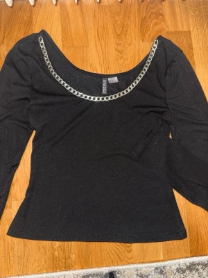 Svart topp med silverkedja från H&M - Snygg svart långärmad topp från H&M Divided med en chunky silverfärgad kedjedetalj längs den breda ringningen. Toppen har en figurnära passform och är tillverkad i mjukt material som känns skönt mot huden. Perfekt för dig som vill sticka ut lite extra.