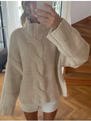 Beige kabelstickad tröja - Säljer en beige stickad tröja med grov kabelstickning framtill och hög krage. Tröjan har breda ärmar och ribbade muddar. Perfekt för dig som gillar en avslappnad och trendig look. 