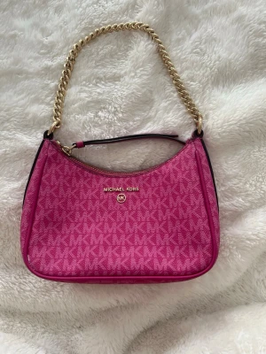 Rosa Michael Kors handväska - Snygg rosa handväska från Michael Kors med guldfärgad kedjerem och dragkedja. Väskan har ett diskret MK-mönster över hela ytan och logotypen i guld framtill. Materialet är tåligt konstläder och modellen är kompakt och stilren, perfekt för att lyfta din outfit.