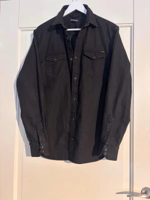 Svart skjorta från Jack & Jones - Snygg svart skjorta från Jack & Jones med klassisk krage, två bröstfickor med knappar och långa ärmar. Skjortan har knappstängning framtill och är tillverkad i ett mjukt bomullstyg. Perfekt för en stilren look.