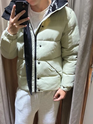 Vit Moncler Jacka - Säljer en vit pufferjacka från Moncler med svart huva och detaljer. Jackan har en quiltad design, dragkedja och tryckknappar framtill samt Moncler-logga på ärmen. Perfekt för kalla vinterdagar och riktigt snygg att bära till jeans eller joggers.