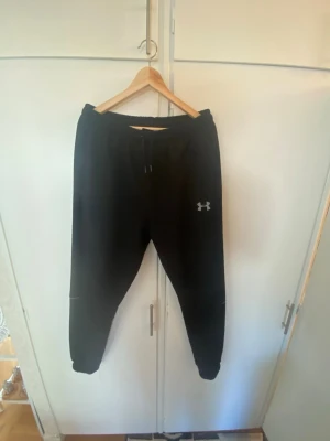 Svarta mjukisbyxor från Under Armour - Säljer ett par svarta mjukisbyxor från Under Armour med dragsko i midjan och smal passform. Byxorna har sidofickor och en liten logga på låret. Perfekta för chill eller träning, riktigt sköna och stilrena.