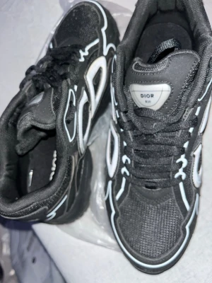 Svarta Dior b30 sneakers med vita detaljer - Snygga svarta Dior sneakers med vita detaljer. Skorna har mesh och syntetmaterial i ovandelen, svarta skosnören och en chunky sula. Märkeslogga på plösen och cool design som sticker ut. Perfekta för dig som vill ha en trendig look. Skorna passar från 43-44