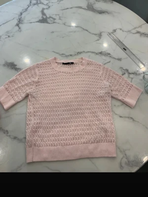 Ljusrosa stickad topp med kort ärm - Söt ljusrosa stickad topp med korta ärmar och rund hals. Tröjan har ett genombrutet mönster och ribbade muddar vid ärmslut och nederkant. Perfekt för lager på lager och ger en fräsch look till din outfit.