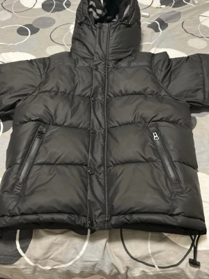 Svart pufferjacka med huva - Säljer en svart pufferjacka med dragkedja och knappar framtill. Jackan har två snedställda fickor med dragkedja och en stor, varm huva. Materialet är glansigt och quiltat, perfekt för kalla vinterdagar. Insidan av huvan har ett rutigt foder i grått och svart.