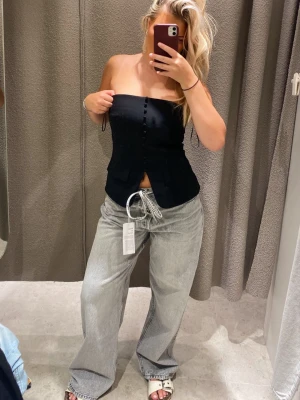 Svart bandeau topp med knappar - Säljer en svart bandeau topp med knappar framtill. Toppen är axelbandslös och har en figurnära passform som ger en snygg siluett. Perfekt att styla med både jeans och kjol för en trendig look.