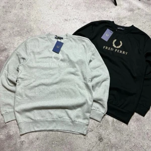 Fred Perry sweatshirt grå & svart - Två snygga sweatshirts från Fred Perry, en i ljusgrått med vitt broderat märke och en i svart med guldigt broderat märke. Klassisk rund halsringning och ribbade muddar. Perfekt för en stilren och avslappnad look.