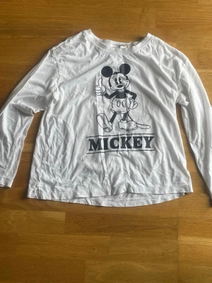 Vit långärmad Mickey Mouse-tröja H&M - Vit långärmad topp från H&M med stort svart tryck av Mickey Mouse och texten 'MICKEY' på framsidan. Tillverkad i mjuk bomull, perfekt för dig som gillar Disney och vill ha en chill och lekfull stil.