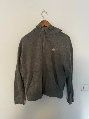 Grå zip hoodie från Nike - Klassisk grå hoodie från Nike med dragkedja framtill och två fickor. Tröjan har huva och Nike-logga broderad på bröstet. Tillverkad i mjukt sweatshirtmaterial som är perfekt för en avslappnad stil.