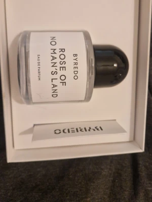 Byredo Rose of No Man's Land parfym - Byredo Rose of No Man's Land Eau de Parfum i en stilren, genomskinlig glasflaska med svart rund kork. Doften är elegant och modern, och flaskan har en vit etikett med svart text. Perfekt för dig som gillar exklusiva och unika parfymer.