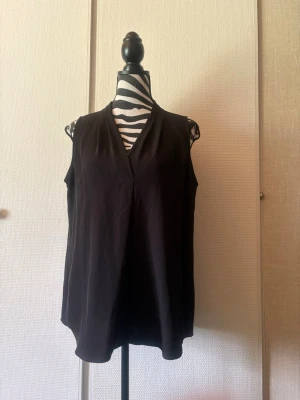 Svart ärmlös v-ringad topp - Stilren svart ärmlös topp med v-ringning och lös passform. Perfekt att matcha med jeans eller kjol för en clean look. Materialet känns mjukt och lätt, vilket gör toppen skön att bära under varma dagar.
