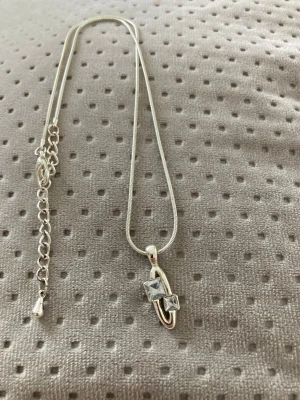 Silverhalsband med fyrkantiga stenar - Ett stilrent silverhalsband med en tunn kedja och ett hängsmycke med två klara, fyrkantiga stenar. Kedjan har justerbar längd med förlängningskedja. Perfekt för att ge en elegant touch till din outfit. Ej äkta silver.