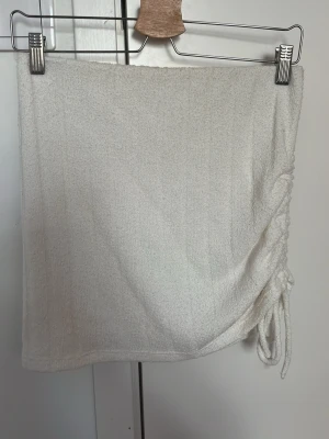 Vit kortkjol med snörning i frotté - Supersnygg vit kortkjol i mjuk frotté med trendig snörning på sidan. Kjolen har en enkel och clean look som passar perfekt till sommaren. Lätt att matcha med olika toppar för en fräsch outfit.