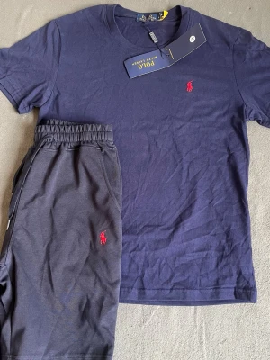 Mörkblått set från Polo Ralph Lauren - Snyggt mörkblått set från Polo Ralph Lauren med t-shirt och shorts. Båda plaggen har det klassiska röda Polo-logot på bröstet och benet. T-shirten har rund hals och shortsen har resårmidja med dragsko. Perfekt för chill dagar eller som loungewear.