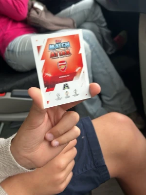 Match Attax Arsenal samlarkort - En  Match Attax samlarkort från Topps med Arsenal-tema. Ett kort visar klubbens logga och det andra är ett spelarkort med målvakten David Raya i gul matchdräkt. Perfekt för dig som samlar på fotbollskort eller gillar Arsenal.