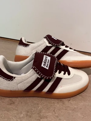 Adidas Samba burgundy och vit - Snygga Adidas Samba sneakers i vitt och burgundy med klassiska tre ränder på sidan. Skorna har en unik detalj med kontrastsöm på tungan och hälen, samt bruna skosnören och gummisula. Materialet är en mix av läder och mocka.