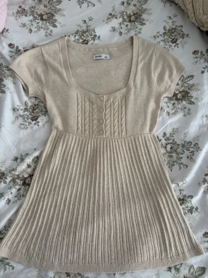 Babydoll top från hollister! - Supersöt beige stickad topp från Hollister med korta ärmar och rundad urringning. Framsidan har dekorativa flätstickade detaljer och fyra knappar. Kjoldelen är ribbstickad för en snygg siluett. Perfekt för dig som gillar en mjuk och stilren look.Skickar inom 1-2 dagar!❤️(har aldrig använt den).