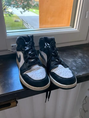 Nike Air Jordan 1 High svart/vit - Säljer ett par Nike Air Jordan 1 High sneakers i svart och vitt läder. Klassisk siluett med svart swoosh, vita paneler och svarta detaljer. Högt skaft och perforerad tåbox för extra komfort. Perfekta för dig som gillar streetwear och ikoniska sneakers.