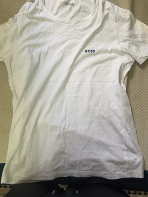 Vit t-shirt från BOSS i bomull - Klassisk vit t-shirt från BOSS med rund halsringning och diskret BOSS-logga på bröstet. Tillverkad i mjuk bomull och har en normal passform. Perfekt basplagg för en clean och stilren look.