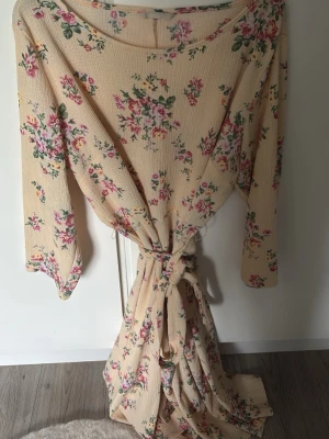 Beige blommig klänning  med knytband - Lång klänning i beige med blommigt mönster i rosa och grönt. Klänningen  har vida ärmar och ett brett knytband i midjan för en avslappnad och romantisk look. Tillverkad i ett mjukt, lätt tyg som känns skönt mot huden.