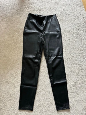 Svarta tights i fuskskinn - Säljer ett par svarta tights i glansigt fuskskinn med hög midja och smal passform. De har en stilren look utan synliga fickor eller detaljer, perfekta för att lyfta vilken outfit som helst.