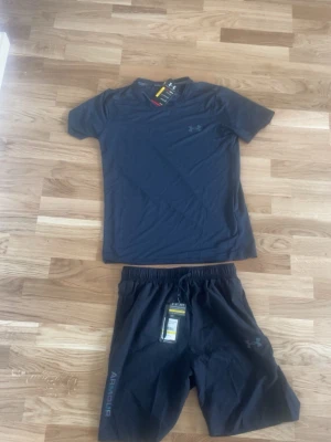 Under Armour träningsset svart - Snyggt träningsset från Under Armour med svart t-shirt och matchande shorts. T-shirten har rund hals och korta ärmar, medan shortsen har resår i midjan och logga på benet. Perfekt för gymmet eller löpning. Tillverkad i lätt och snabbtorkande material.