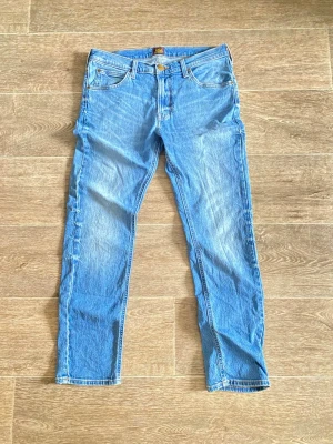 Blå raka jeans från Lee - Säljer ett par klassiska blå jeans från Lee med rak passform och fem fickor. Jeansen har en ljus tvätt och Lee-lapp bak i midjan. Tillverkade i slitstark denim, perfekta för en avslappnad och tidlös stil.
