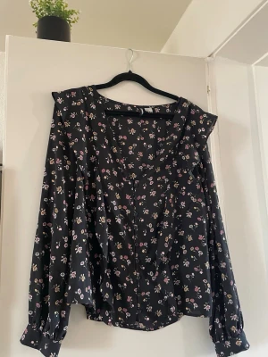 Blommig svart blus från Divided - Snygg svart blus från Divided med små blommor i rosa, gult och vitt. Blusen har långa ärmar med breda manschetter och knappar framtill. Tillverkad i ett lätt och luftigt material som ger en avslappnad men trendig look.
