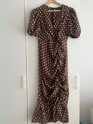 Brun prickig långklänning med puffärm - Snygg brun långklänning med vita prickar och puffärmar. Klänningen har v-ringning, rynkade detaljer framtill och volang längst ner. Perfekt för dig som vill sticka ut med ett retroinspirerat plagg.