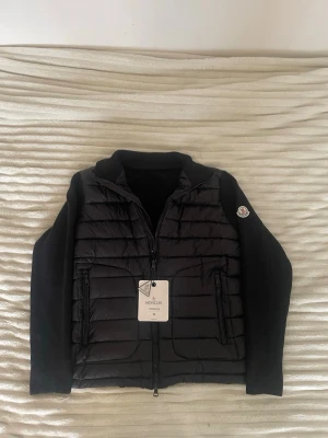 Moncler cardigan  - Säljer en helt ny och oanvänd Moncler hybridcardigan i svart! Stickade ärmar med dunjacka-front. Alla taggar sitter kvar (10/10 skick).  Storlek: M på lappen, men Moncler är små i storleken så den passar perfekt som en Small.  NFC-taggen fungerar och scannas direkt till moncler.com med mobilen. QR-kod och hologram finns på tvättlappen på insidan. Kan fraktas spårbart! Hör av dig vid frågor ✨ 