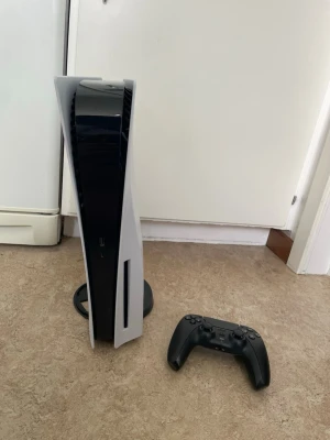 PlayStation 5 med svart handkontroll - Säljer en PlayStation 5 konsol från Sony med futuristisk design i vitt och svart. Konsolen har en slimmad, stående form och medföljer gör en svart DualSense handkontroll. Perfekt för gaming och streaming av högsta kvalitet.