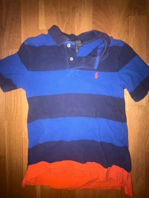 pikétröja från Polo Ralph Lauren - Snygg pikétröja med breda marinblå och blå ränder samt orange detalj längst ner. Klassisk krage med knappar och den ikoniska Polo-loggan broderad i orange på bröstet. Perfekt för en avslappnad men ändå stilren look.