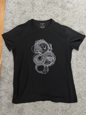 Svart t-shirt med draktryck från SHEIN - Cool svart t-shirt från SHEIN med ett detaljerat vitt drakmotiv på framsidan. Klassisk rund hals och korta ärmar. Materialet är mjukt och känns som bomull eller polyester. Perfekt för dig som gillar edgy prints och vill sticka ut.