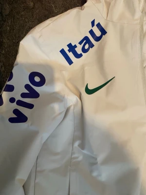 White windbreaker Brazil - Snygg vit vindjacka från Nike med Brasilien-tema. Jackan har blåa tryck med 'Itaú' och 'vivo' samt en grön Nike-logga på bröstet. Tillverkad i lätt och tåligt material, perfekt för blåsiga dagar. Dragkedja framtill och huva för extra skydd. Passformen är M/S men det står L