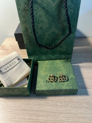 Guldiga GG örhängen från Gucci - Säljer ett par ikoniska örhängen från Gucci i guldfärgad metall med det klassiska dubbla G-logotypen. Örhängena har en stilren och tidlös design som passar perfekt till alla outfits. Levereras i originalask och med tygpåse från Gucci.
