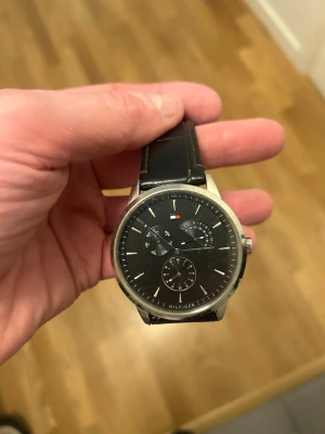 Svart klocka från Tommy Hilfiger - Stilren klocka från Tommy Hilfiger med svart urtavla och silverfärgat boett. Klockan har flera små urtavlor och svart läderarmband. Perfekt accessoar för dig som gillar klassisk och tidlös design.