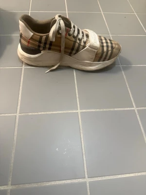 Burberry sneakers med klassiskt rutmönster - Snygga sneakers från Burberry med det ikoniska beige, svarta och röda rutmönstret. Skorna har vita snören, bred vit rem med Burberry-logga och en chunky vit sula. Materialet är en mix av textil och läderdetaljer. Perfekta för dig som gillar lyxig streetstyle. Priset går o sänka vid en snabbare äfär möts upp i Göteborg eller så går det o frakta igenom plick