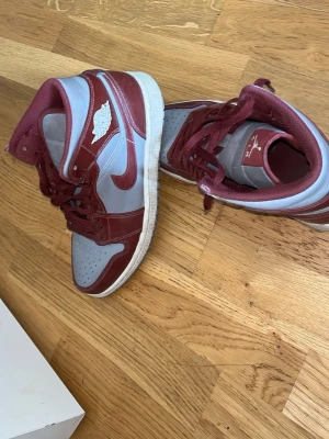 Air Jordan 1 Mid Bordeaux/Grå - Snygga Air Jordan 1 Mid sneakers i bordeauxrött och ljusgrått. Klassisk siluett med vita detaljer och ikoniska vingsloggan på sidan. Skorna har en kombination av läder och syntetmaterial samt vit sula. Perfekta för dig som gillar streetwear och vill sticka ut.