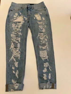 Ljusblå slitna jeans med hål Tiger of Sweden  - Säljer ett par ljusblå jeans med extremt slitna detaljer och stora hål längs båda benen. Jeansen har klassisk femficksmodell, uppvikta benslut och är gjorda i denim. Perfekta för dig som gillar en edgy och trendig stil.
