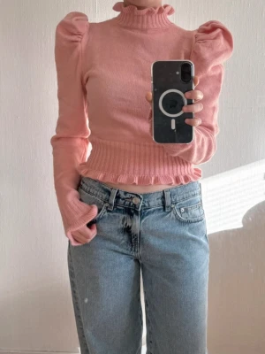 Rosa topp med puffärmar och volang från Cider - Söt rosa tröja med puffärmar och volangdetaljer vid hals, midja och ärmslut. Strl XS. Stickad och har en hög hals med ribbad kant. Från Cider. Oanvänd med prislapp kvar. 