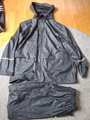 Svart regnställ från Bluewear M - Säljer ett svart regnställ från Bluewear i storlek M. Jackan har huva, dragkedja med tryckknappar, två stora fickor och reflexdetaljer på ärmarna. Materialet är vattenavvisande och byxorna har resår i midjan för skön passform.