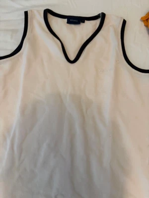 Vit linnetopp med svarta kanter Redgreen - Säljer en vit linnetopp från Redgreen med svarta kontrastkanter och v-ringad hals. Toppen är ärmlös och har en stilren, sportig look som passar perfekt till sommaren. Materialet känns som bomull och toppen har en diskret logga på bröstet.