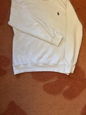 Vit sweatshirt från Ralph Lauren - Klassisk vit sweatshirt från Ralph Lauren med rund halsringning och den ikoniska mörkblå polospelaren broderad på bröstet. Tröjan har ribbade muddar vid ärmslut och nederkant. Perfekt för en clean och stilren look.