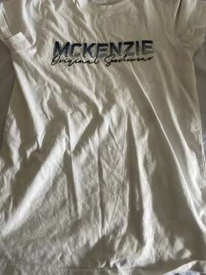 Vit McKenzie t-shirt med tryck - Säljer en vit t-shirt från McKenzie med blått och svart tryck på bröstet där det står 'McKenzie Original Sportswear'. Klassisk rund halsringning och korta ärmar. Perfekt till jeans eller shorts för en avslappnad look.