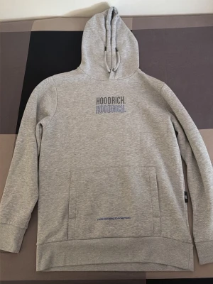 Grå hoodie från Hoodrich - Snygg grå hoodie från Hoodrich med tryck både fram och bak. Klassisk känguruficka och justerbar huva med dragsko. Tillverkad i mjukt bomullsmaterial som är perfekt för chill dagar. Stora loggor och texten 'FROM NOTHING TO SOMETHING' ger streetwear-vibe.