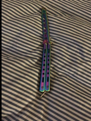 Regnbågsfärgad butterfly-träningskniv - Säljer en butterfly-träningskniv i metall med regnbågsfärgad finish. Handtaget har flera hål och cool design, perfekt för att öva tricks och flips. Ingen skarp egg, så den är gjord för träning och tricks. Snygg accessoar för dig som gillar att samla eller träna.
