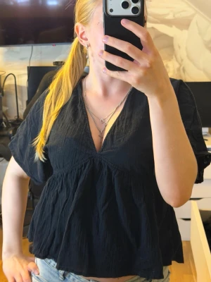 Svart v-ringad blus med kort ärm - Säljer en svart blus med v-ringning och avslappnad passform. Blusen har korta ärmar och är gjord i ett tunt, lätt och lite skrynkligt tyg som ger en cool och ledig vibe. Perfekt att matcha med jeans eller kjol för en trendig look.