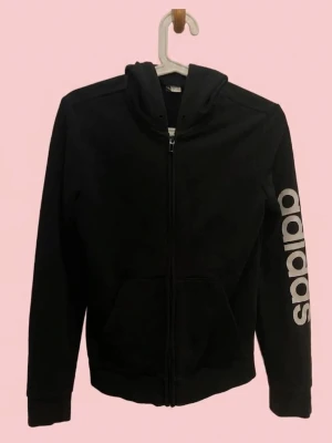 Svart hoodie från Adidas med tryck - Säljer en svart hoodie från Adidas med dragkedja framtill och stor vit Adidas-logga längs ena ärmen. Hoodien har huva och två fickor framtill. Tillverkad i mjukt material som passar perfekt till en avslappnad stil.