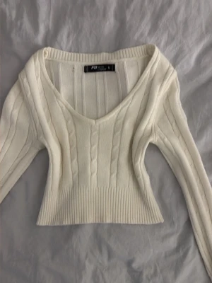 Vit kabelstickad croppad tröja - Säljer en vit kabelstickad tröja från PrettyLittleThing. Tröjan har croppad passform, djup v-ringning och långa ärmar. Ribbstickad kant nedtill och vid ärmslut. Perfekt för lager på lager och en clean look.